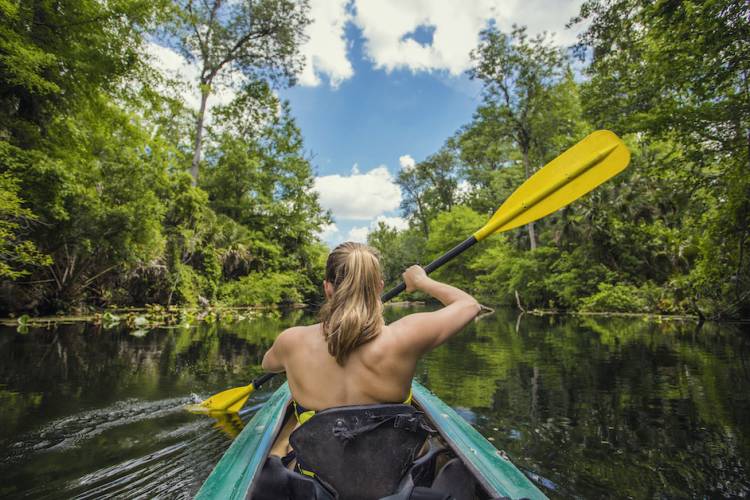 woman kayaking river with view from behind her as she goes through narrow passage in trees